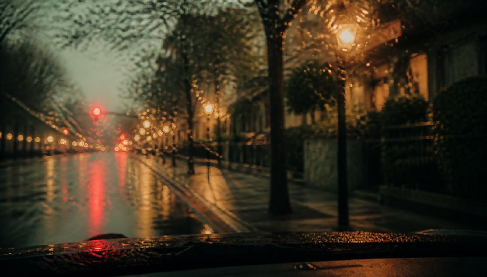 Rainy city street at dusk seen through a windshield, red taillights reflecting on wet asphalt